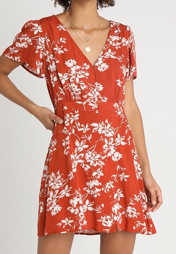 Femme portant une robe orange rouille à manches courtes et col en V, imprimée de fleurs blanches, avec des colliers en or superposés, posant avec une main sur la hanche.