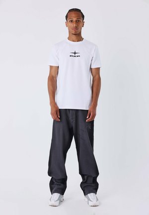 T-shirt bianco a maniche corte con logo "PXP" nero, abbinato a pantaloni grigi scuri dalla vestibilità ampia e sneakers bianche. Design semplice e pulito.