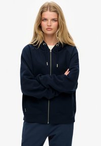 Sudadera azul marino con cremallera, capucha con cordón, grandes bolsillos frontales y puños acanalados. Tejido suave con una textura lisa.
