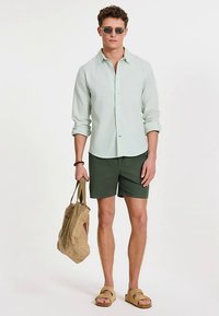 Svijetlozelena košulja s dugim rukavima na kopčanje, tamnozelene kratke hlače, bež sandale i bež torba za nošenje, dodatno ukrašeni sunčanim naočalama.