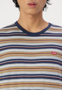 Un jeune portant une chemise Levi's rayée marron, blanche et bleu marine avec un col rond et un patch logo Levi's rouge sur la poitrine.