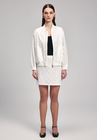 Giacca bomber bianca cropped con dettagli ad occhio, abbinata a una maglietta nera e una mini gonna bianca con occhielli. Tacchi neri con accenti dorati.