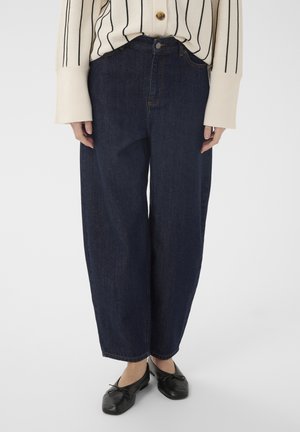 Personne portant un jean large bleu marine, un pull crème à rayures noires et des ballerines noires, debout devant un fond uni.