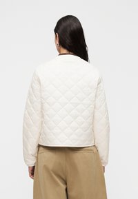 Καπιτονέ, εκρού μπουφάν με κοντή σχεδίαση και στρογγυλή λαιμόκοψη. Διαθέτει καφέ αντιθετικές λεπτομέρειες και μακριά μανίκια.