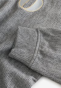 Detail sivého pleteného rukáva svetra zobrazujúci textúrovanú tkaninu a malý vyšívaný vzor so žltou a sivou niťou.