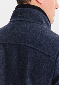 Mężczyzna w ciemnoniebieskim, teksturowanym płaszczu z podniesionym kołnierzem, ukazujący tył i profil boczny jego głowy i szyi.