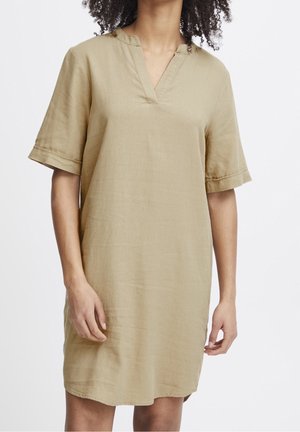 Femme portant une robe beige ample à manches courtes avec un col en V et sans col, debout devant un fond blanc uni.