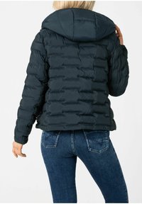 Dunkelblaue Steppjacke mit horizontaler Steppoptik, Kapuze und glatter Textur. Getragen über blauen Jeans mit sichtbaren Steppdetails.