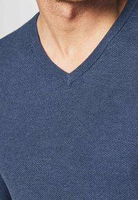 Blauer gestrickter V-Ausschnitt-Pullover, der das strukturierte Gewebe zeigt, mit einer Nahaufnahme des Halsausschnitts und des oberen Brustbereichs.