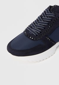 Ναυτικό μπλε αθλητικό παπούτσι με λευκά κορδόνια σε σχήμα ζιγκ-ζαγκ, μαύρες λεπτομέρειες με στρας, σουέτ διακοσμήσεις και παχιά  λευκή υφασμένη σόλα.