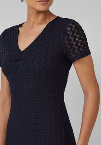 Robe marine avec un décolleté en V et des manches courtes, mettant en avant un motif en tricot texturé et légèrement froncé au niveau de la poitrine.
