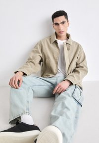 Beige kordfløyelsjakke med trykknapper og lommer, kombinert med lyseblå denimbukser med glidelåslomme. Svarte joggesko med hvit såle.