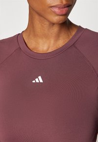 Burgundijska sportska majica s okruglim izrezom i bijelim logom, izrađena od glatkog, rastezljivog materijala s raglan rukavima.