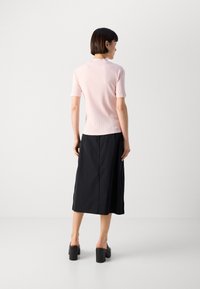 Helles rosa, geripptes T-Shirt mit kurzen Ärmeln, kombiniert mit einem langen schwarzen Rock. Das Modell trägt schwarze Blockabsatzmules. Einfaches, modernes Design.
