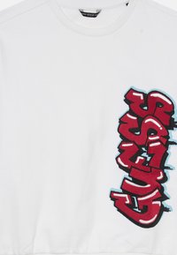 Fehér crew neck pulóver, jobboldalt függőleges piros graffiti-stílusú "GUESS" logóval, fekete és kék körvonalazással.