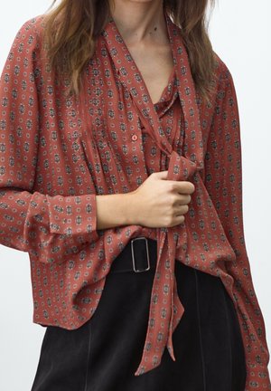 Woman wearing rust-colored patterned blouse with tie neck detail and black high-waisted pants with a square buckle belt.