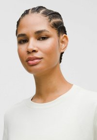 Vrouw met gevlochten haar, gekleed in een witte crewneck top, die iets opzij kijkt tegen een lichte effen achtergrond.