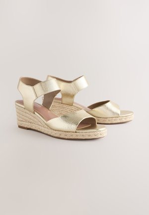 Kuldse metallilise viimistlusega kiilkontsaga sandaalid kahekordsete rihmadega, avatud varbaga ja punutud nöörpõhjaga. Siledapinnaline ja läikiva viimistlusega.