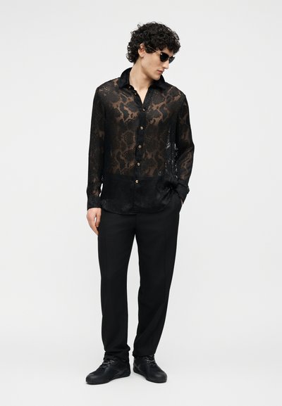 Homme aux cheveux bouclés portant des lunettes de soleil noires, une chemise noire transparente à motifs, un pantalon noir et des baskets noires, debout devant un fond blanc.