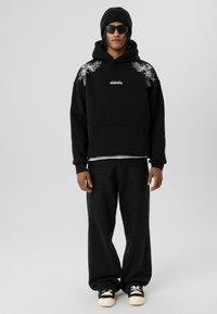 Zwarte hoodie met witte bloemenborduursels op de schouders, voorzak, gedragen over een wit shirt, gecombineerd met zwarte wijde jeans en zwarte sneakers.