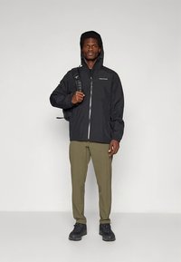 Schwarze wasserdichte Jacke mit Kapuze, Frontreißverschluss und Logo. Dazu olivgrüne Hose und schwarze Stiefel getragen. Rucksack über eine Schulter gehängt.