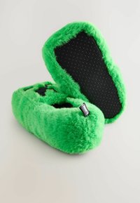 Chaussons en peluche vert vif avec des semelles antidérapantes noires, un chausson posé à plat et l'autre incliné verticalement.