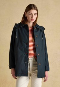 Młoda kobieta w ciemnoniebieskim płaszczu zapinanym na guziki, na czerwono-białym, paskowanym swetrze i jasnobeżowych spodniach na jednolitym beżowym tle.