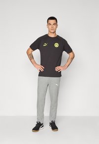 Sort kortærmet t-shirt med gule logoer, parret med lys grå sweatpants og sorte sneakers, stående i en neutral positur.