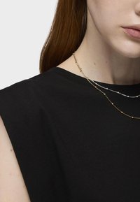 Dos collares superpuestos: uno plateado con pequeñas cuentas, y uno dorado con acentos redondeados espaciados, llevados sobre una blusa de tela negra.