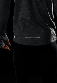Maglietta atletica grigia a maniche lunghe con tessuto testurizzato e una striscia riflettente bianca all'orlo, abbinata a leggings neri aderenti.