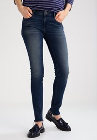 Jeans ajustados de color azul con un lavado desgastado, cinturilla alta y diseño clásico de cinco bolsillos. Combinados con mocasines oscuros para un contraste.