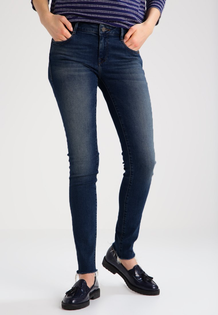 Jeans ajustados de color azul con un lavado desgastado, cinturilla alta y diseño clásico de cinco bolsillos. Combinados con mocasines oscuros para un contraste.
