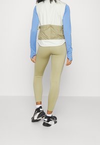 Chaleco ligero a bloques de color en blanco, azul y caqui, combinado con leggings verde oliva y zapatillas deportivas negras con acentos blancos.