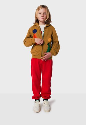Sudadera con cremallera marrón con diseños geométricos coloridos, pantalones de chándal rojos y zapatillas blancas. Tejido suave con un corte relajado.