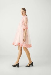 Kvinna i en ljusrosa genomskinlig klänning med blommig detaljer på ärmar och fåll, bärande svarta blockklackade Mary Jane-skor, stående på sidan.