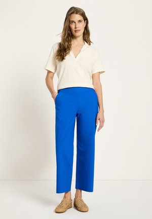 Woman wearing a white short-sleeve blouse, bright blue straight-leg pants, and woven tan slip-on shoes, standing with hands in pockets.