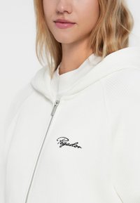 Bijela rebrasta dukserica na zip s prilagođenim kapuljačom i izvezenim "Pegador" logom u crnoj boji na lijevoj strani prsiju. Glatka tekstura i moderni dizajn.