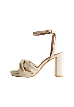 Sandalo beige con tacco alto, cinturino alla caviglia, chiusura con fibbia, cinturino frontale annodato e tacco a blocco su sfondo bianco.