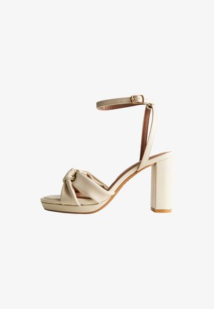 Sandale à talon haut beige avec bride cheville, fermeture à boucle, bride avant nouée et talon bloc sur fond blanc.