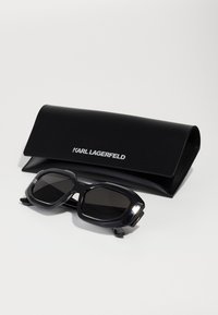 Occhiali da sole neri oversize con montatura geometrica e lenti scure, accompagnati da un elegante astuccio nero con marchio in argento.