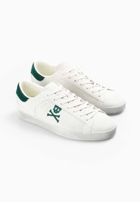 Zapatillas blancas con detalles en verde, que presentan un diseño de calavera y huesos cruzados, material texturizado y suela de goma plana.