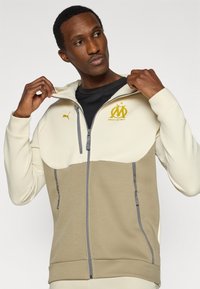 Φωτεινή μπεζ και χακί ζακέτα με φερμουάρ και ψηλό γιακά, με χρυσό λογότυπο και γκρι λεπτομέρειες, κατασκευασμένη από απαλό ύφασμα με άνετη, καθημερινή εφαρμογή.