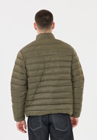 Olivgrön pufferjacka med hög krage, stickade horisontella sömmar och slät yta. Bärs över mörka denimjeans.