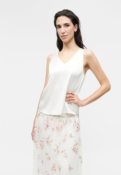 Femme aux cheveux foncés portant un haut en satin blanc sans manches et une jupe blanche transparente avec broderie florale rose, posant avec la main sur la hanche.