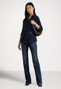 Tumši zila pogājama krekla ar mezgla detaļu, kombinēta ar izceltām džinsu biksēm un apaļu melnu ādas plecu somu ar zelta aizdari.