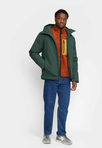 Green waterproof jacket with a hood over an orange fleece, featuring a tan chest pocket. Worn with blue jeans and grey sneakers.