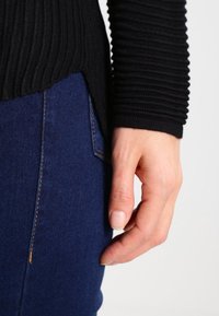 Schwarzer gerippter Strickpullover mit langen Ärmeln, kombiniert mit dunkelblauen Jeans. Nahaufnahme einer Hand, die auf der Jeans mit auffälligen Nähdetails ruht.