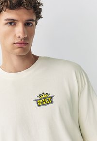 Vaaleabeige puuvillainen t-paita, jossa on pyöreä pääntie ja vasemmassa rintataskussa värikäs "OUTKAST"-logo sekä kruunukuvio.