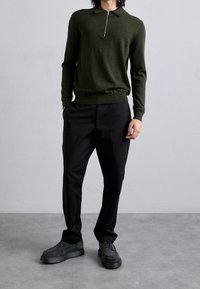 Polo de punto de color verde oscuro con cremallera, combinado con pantalones negros. Calzado con material texturizado negro y suela gruesa.