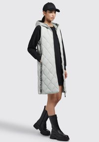 Gepolsterte, hellgraue Weste mit Kapuze, seitlichen Taschen und schwarzem Branding. Über einem schwarzen Kleid getragen, kombiniert mit knielangen, schwarzen Schnürstiefeln.